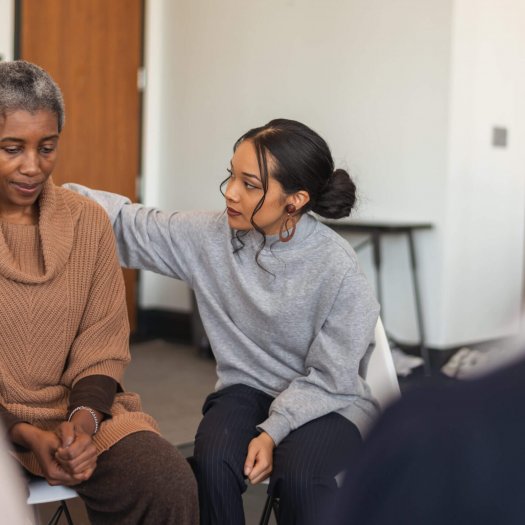 A multi-ethnic group of adults are attending a group therapy session. The attendees are seated in a circle. A senior black woman is sharing her struggles with the group. A mixed-race young woman rests her arm on the woman's back, expressing comfort and support.