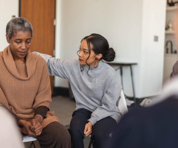 A multi-ethnic group of adults are attending a group therapy session. The attendees are seated in a circle. A senior black woman is sharing her struggles with the group. A mixed-race young woman rests her arm on the woman's back, expressing comfort and support.